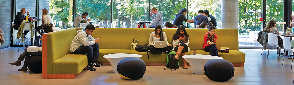 Landscape of U of T students sitting in one of the building lobbies on a couch and at tables talking, working on laptops and using cellphones.