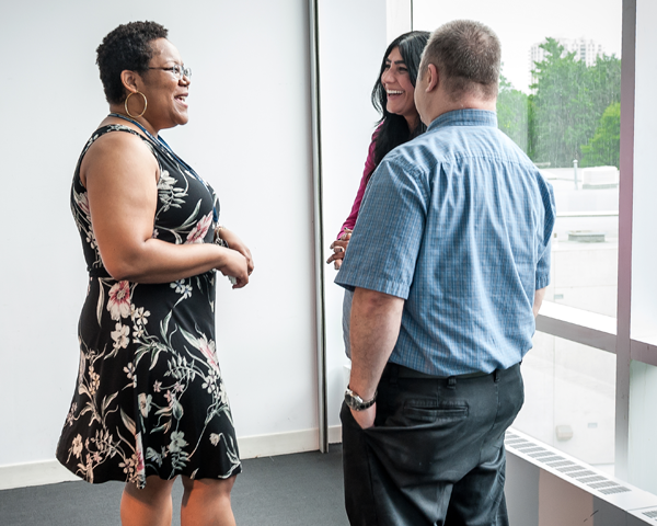 Three U of T staff members standing indoors by a window on campus chatting and laughing.