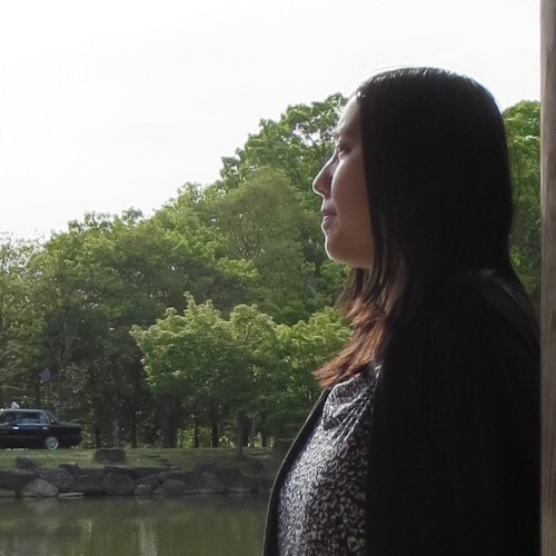 Woman stands in front of forest and lake looking into distance.