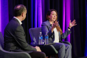 Donna Kidwell sits in chair on stage and gestures with hands in air while Shane Schick, captured from behind, observes.